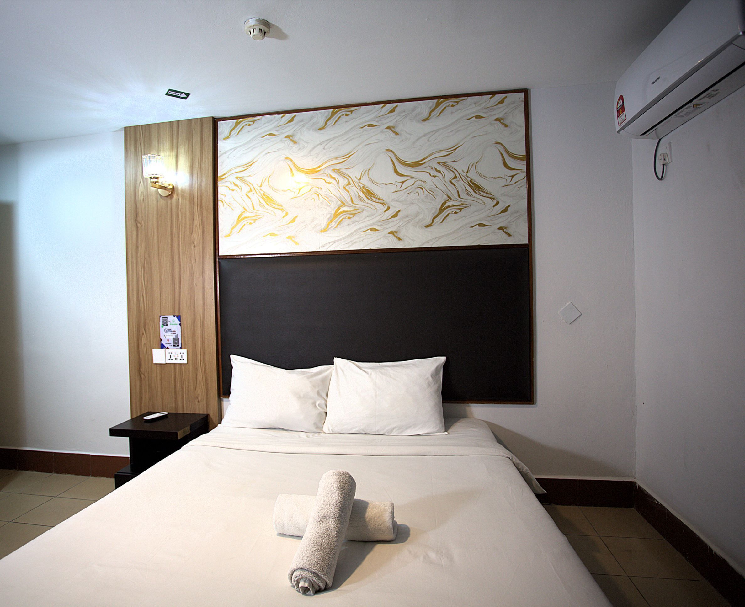 Habitación de hotel con una cama grande, cabecera con diseño tipo mármol, mesita de noche con control remoto y un equipo de aire acondicionado en la pared.
