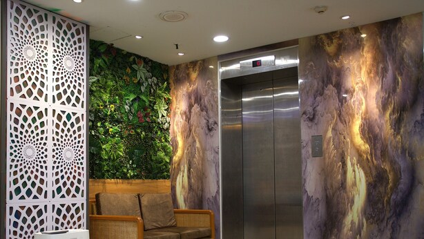 An elevator with a metallic finish, a wall with a green leafy mural, and a wicker sofa with cushions. - Hotel Putra Hills at WTCKL (Kuala Lumpur)
