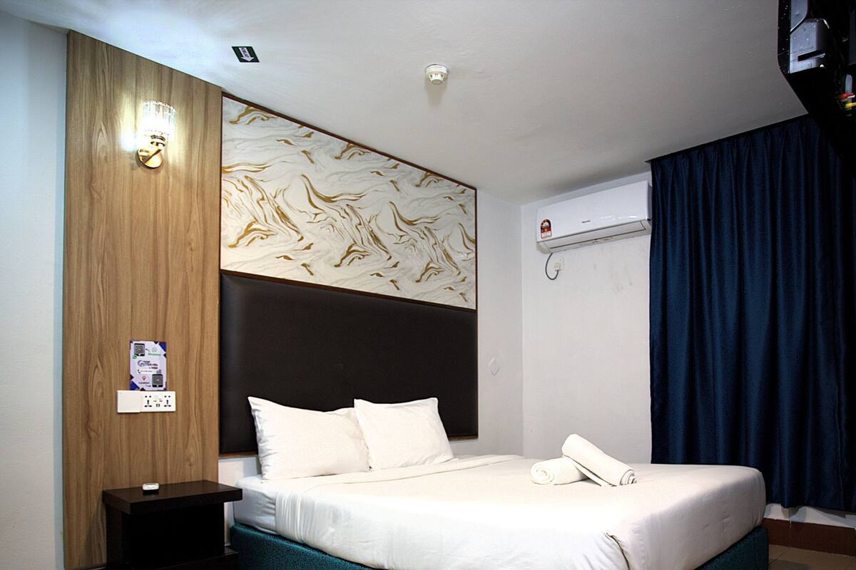 Ein Hotelzimmer mit Bett, Nachttisch, Wandairconditioner und einem mit Vorhängen versehenen Fenster.