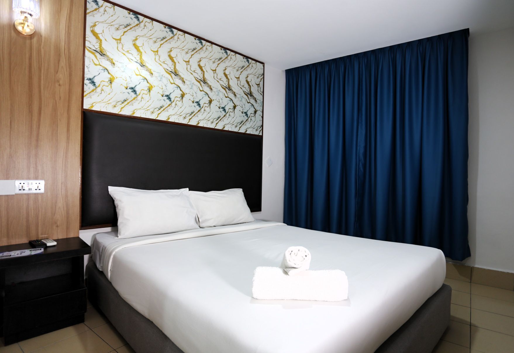 A hotel room with a bed, a nightstand, a wall with a marble-like pattern, and a large window with blue curtains.