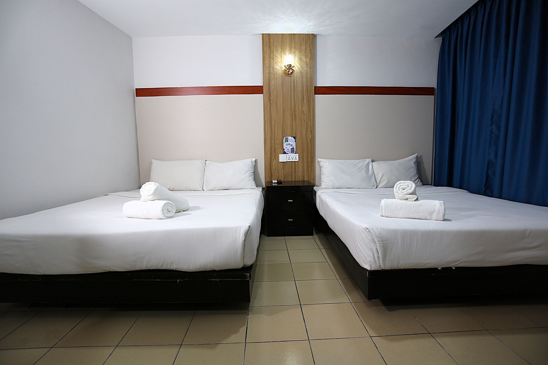 Una habitación de hotel con dos camas individuales, un panel de madera decorativo, una mesita de noche y una cortina azul.