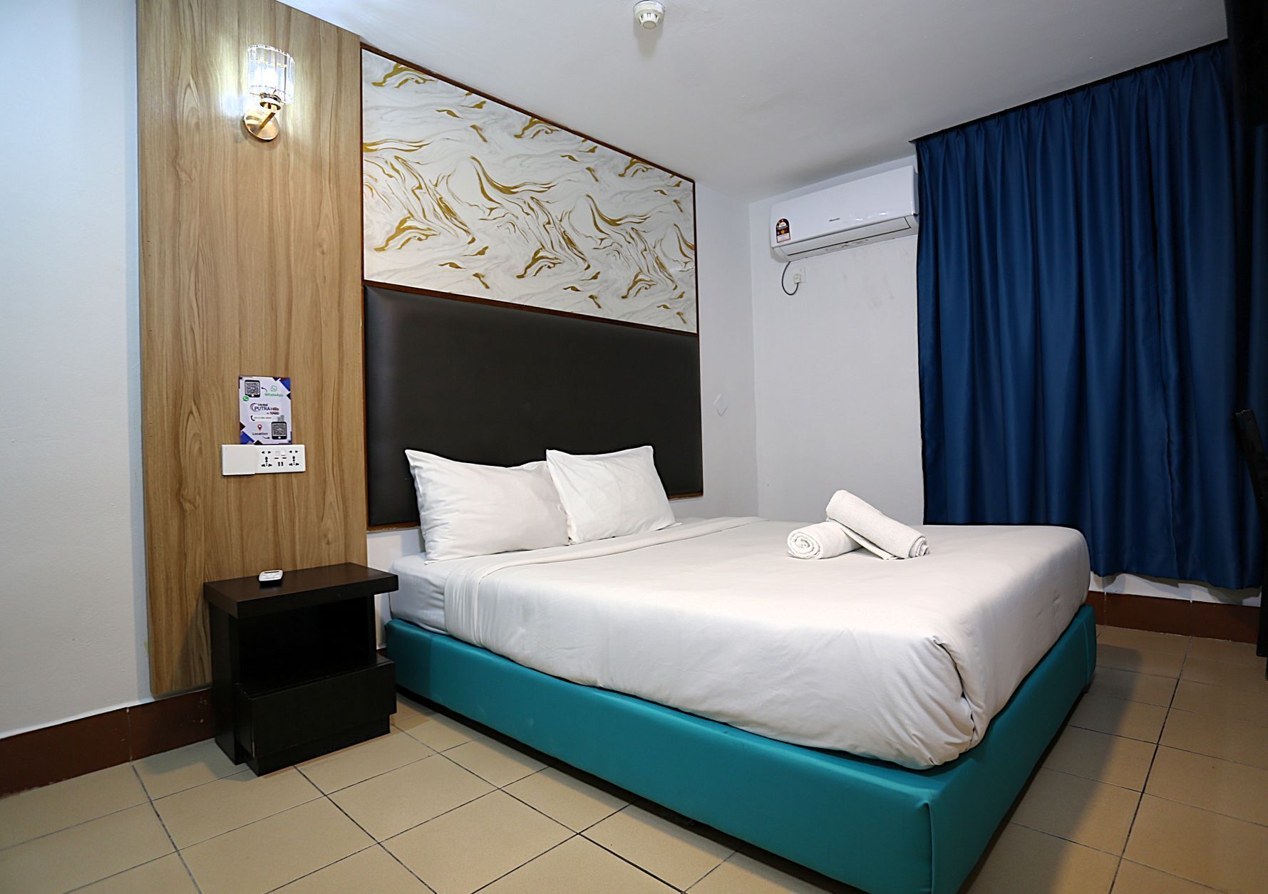 Une chambre d’hôtel avec un lit, une table de chevet, un climatiseur fixé au mur et un rideau bleu.