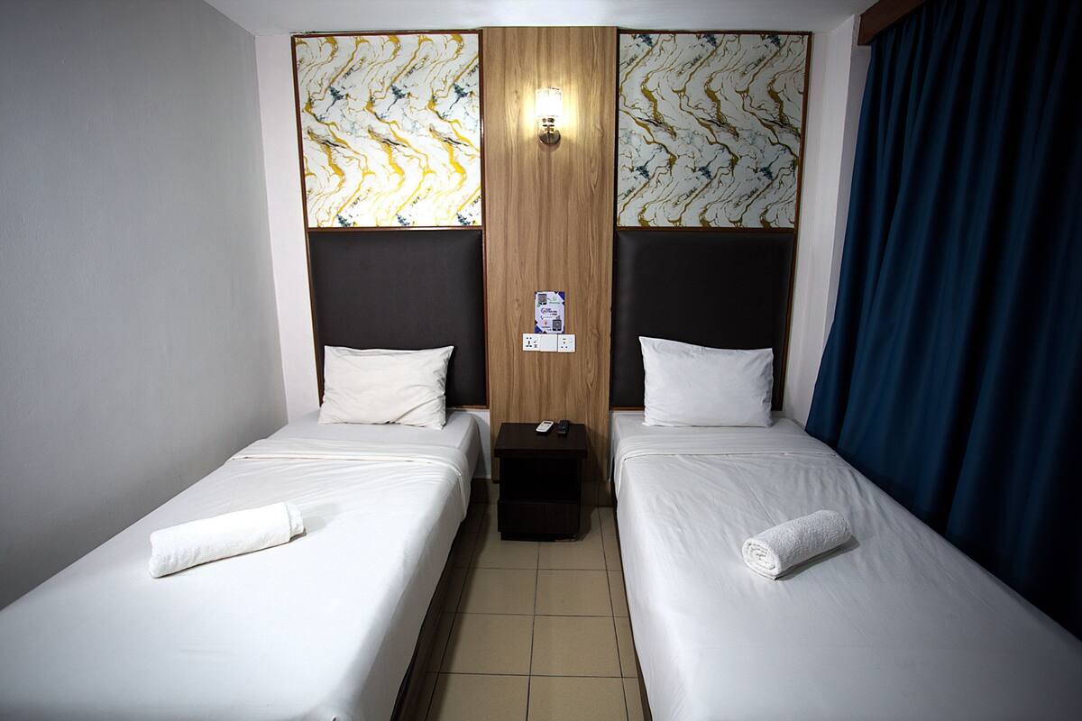Ein Hotelzimmer mit zwei Einzelbetten, einem Holz-Kopfteil, einem Nachttisch mit Fernbedienung und einer Wandleuchte.