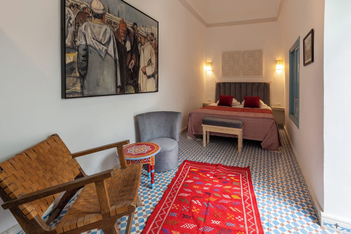 Quarto com cama, cadeira, mesinha e um quadro grande na parede.