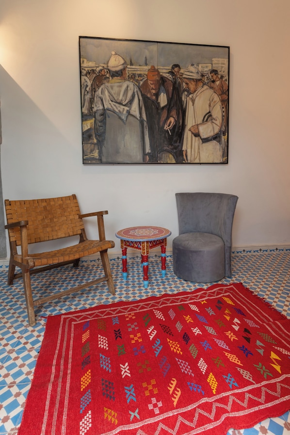 A room with a wooden chair, a grey armchair, a small round table with a colorful patterned tablecloth, and a large painting on the wall. - Palais el Abbassia (Marrakech)