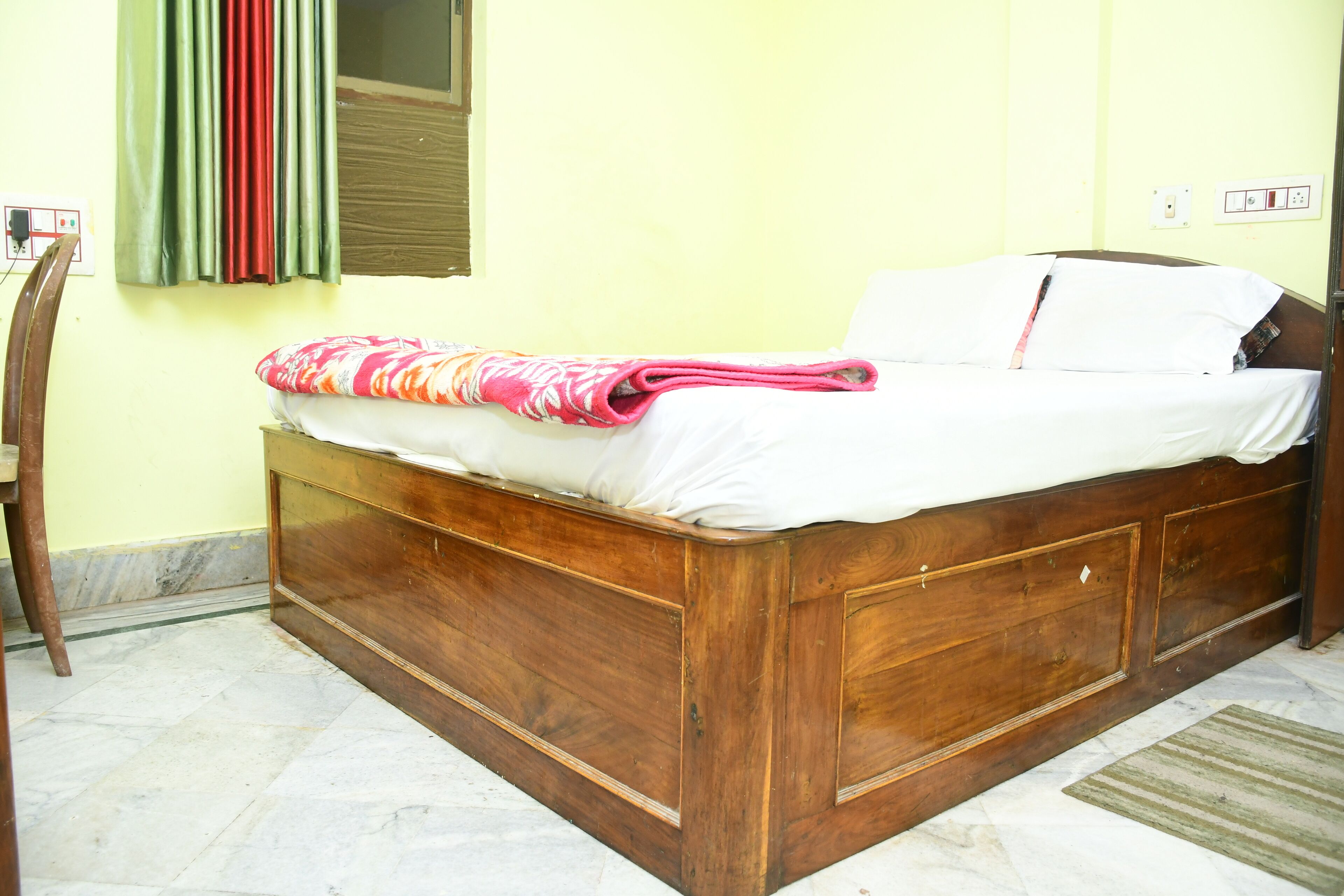 Un lit en bois avec un matelas blanc et une couverture rose, une chaise en bois et une fenêtre ornée de rideaux verts et rouges.