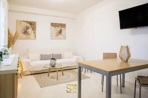 Ein modernes Wohnzimmer mit Sofa, Couchtisch und Esstisch. An den Wänden hängen zwei abstrakte Gemälde, über dem Sofa ist ein Flachbildfernseher angebracht und auf dem Esstisch steht eine Vase.
