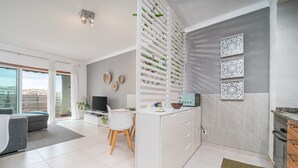 A modern kitchen with a central island, a dining area with a grey sofa and white chairs, and a view of the city through large windows. - Sunny Apartment with Shared Pool (Portimão)