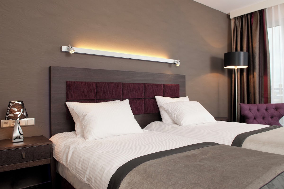 Habitación de hotel con una cama grande, una mesita de noche, una lámpara y vistas a la ciudad desde la ventana.