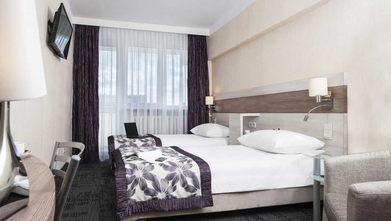 A hotel room with a bed, bedside tables, a desk, a chair, a TV, and a window with curtains.