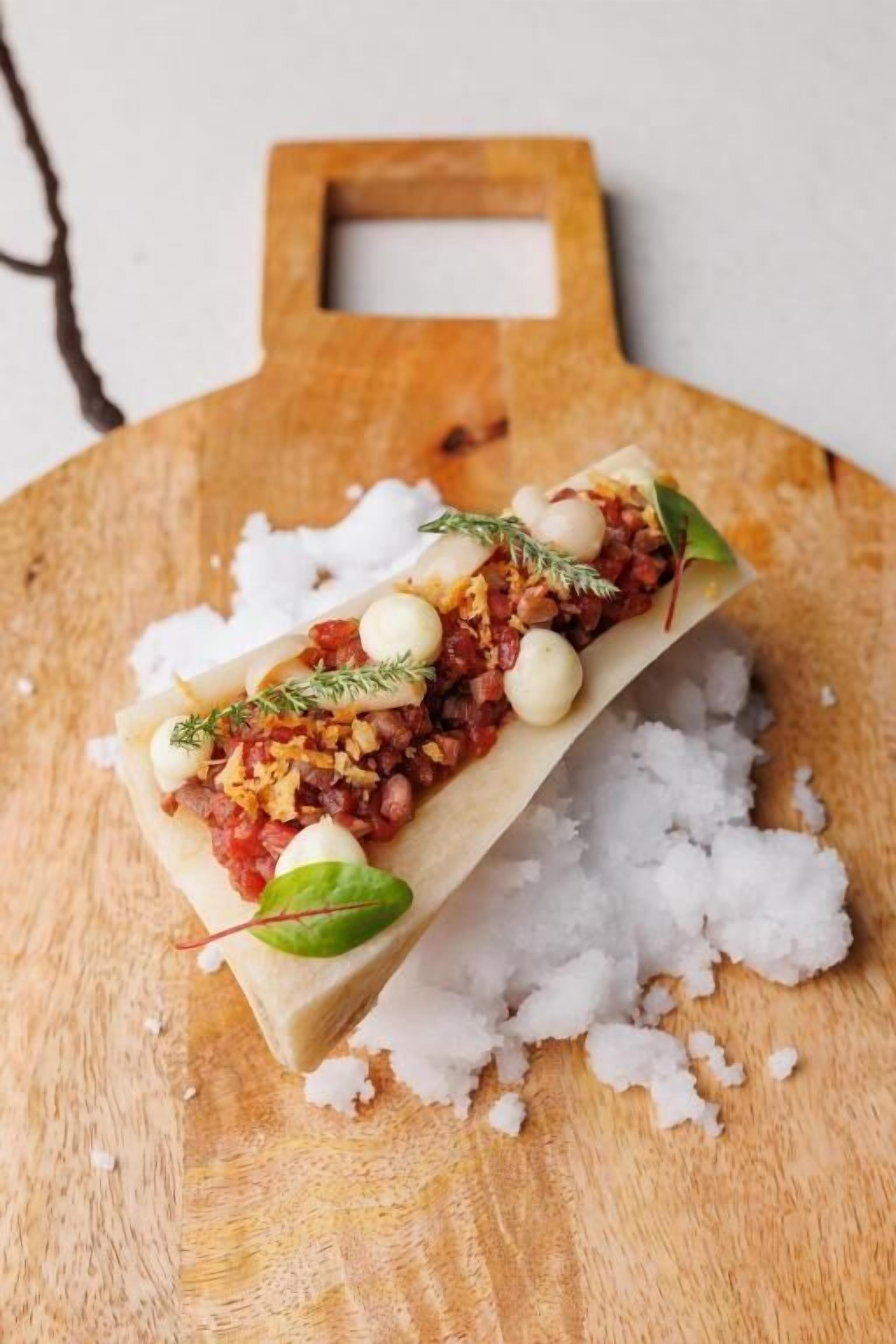 Una taula de fusta amb un plat rectangular que conté un aliment en capes, decorat amb herbes i petites boles rodones, damunt d'una superfície de fusta amb sal escampada.