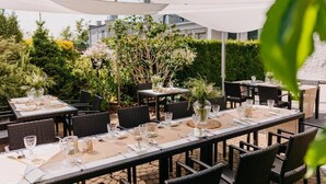 An outdoor dining area with tables set for two, surrounded by greenery and a white building in the background. - Hotel 3Trio (Bialystok)