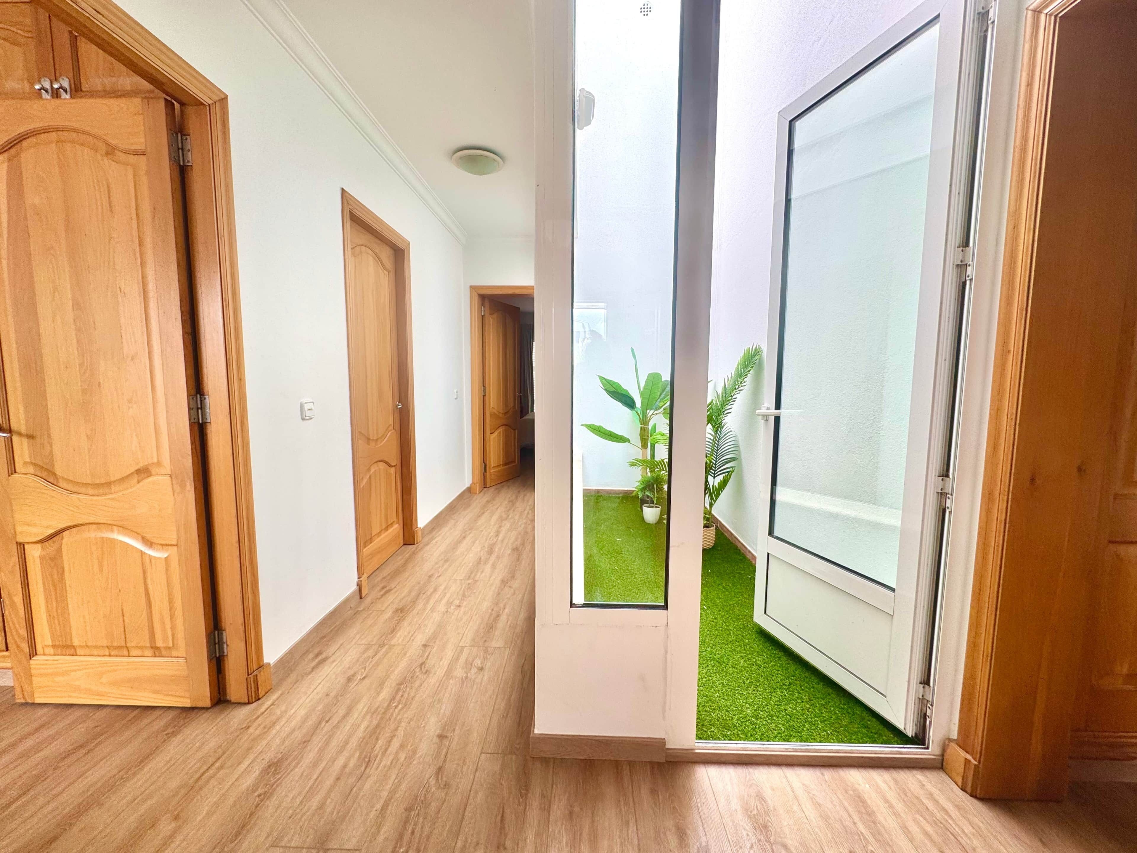 Un pasillo con puertas de madera, una puerta de cristal que da a un balcón con césped artificial y plantas en macetas.