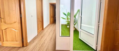 Un pasillo con puertas de madera, una puerta de cristal que da a un balcón con césped artificial y plantas en macetas.