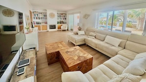 Een ruime zithoek met een grote beige hoekbank, een houten salontafel en een open haard. Er staan boekenkasten vol boeken en decoratieve objecten, en een groot raam biedt uitzicht op de omgeving.
