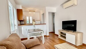 Een moderne woonkamer met een bank, een salontafel, een televisie op een houten standaard, een eethoek met een tafel en stoelen, en een keuken met kastjes en een magnetron.