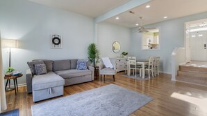 Ein modernes Wohnzimmer mit grauem Sofa, einem kleinen Tisch mit Lampe, einem Essbereich mit weißem Tisch und Stühlen sowie einer Treppe, die eine weitere Ebene hinaufführt.
