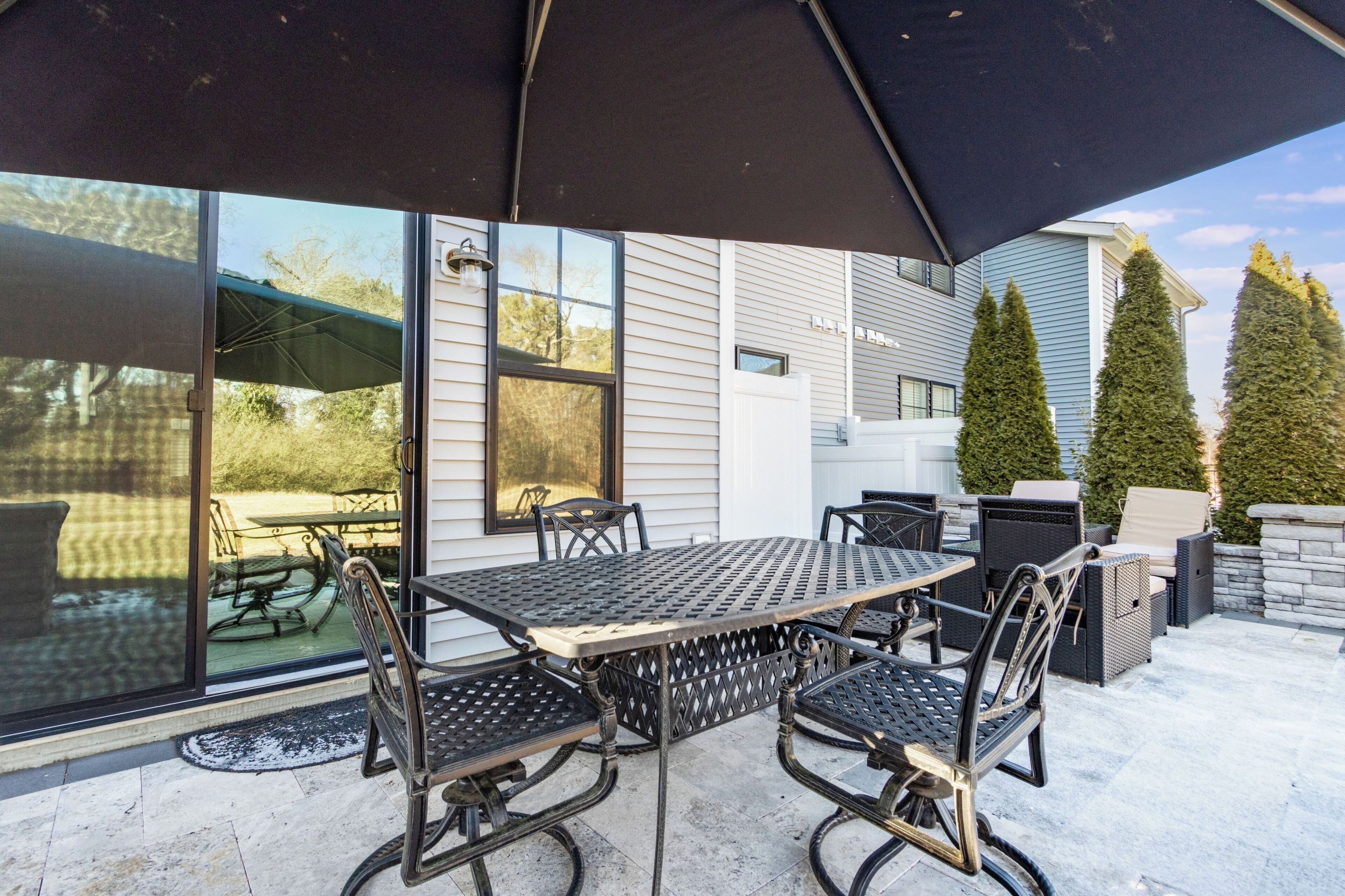 An outdoor patio with a table and chairs, a large umbrella, and a view of a house and trees.