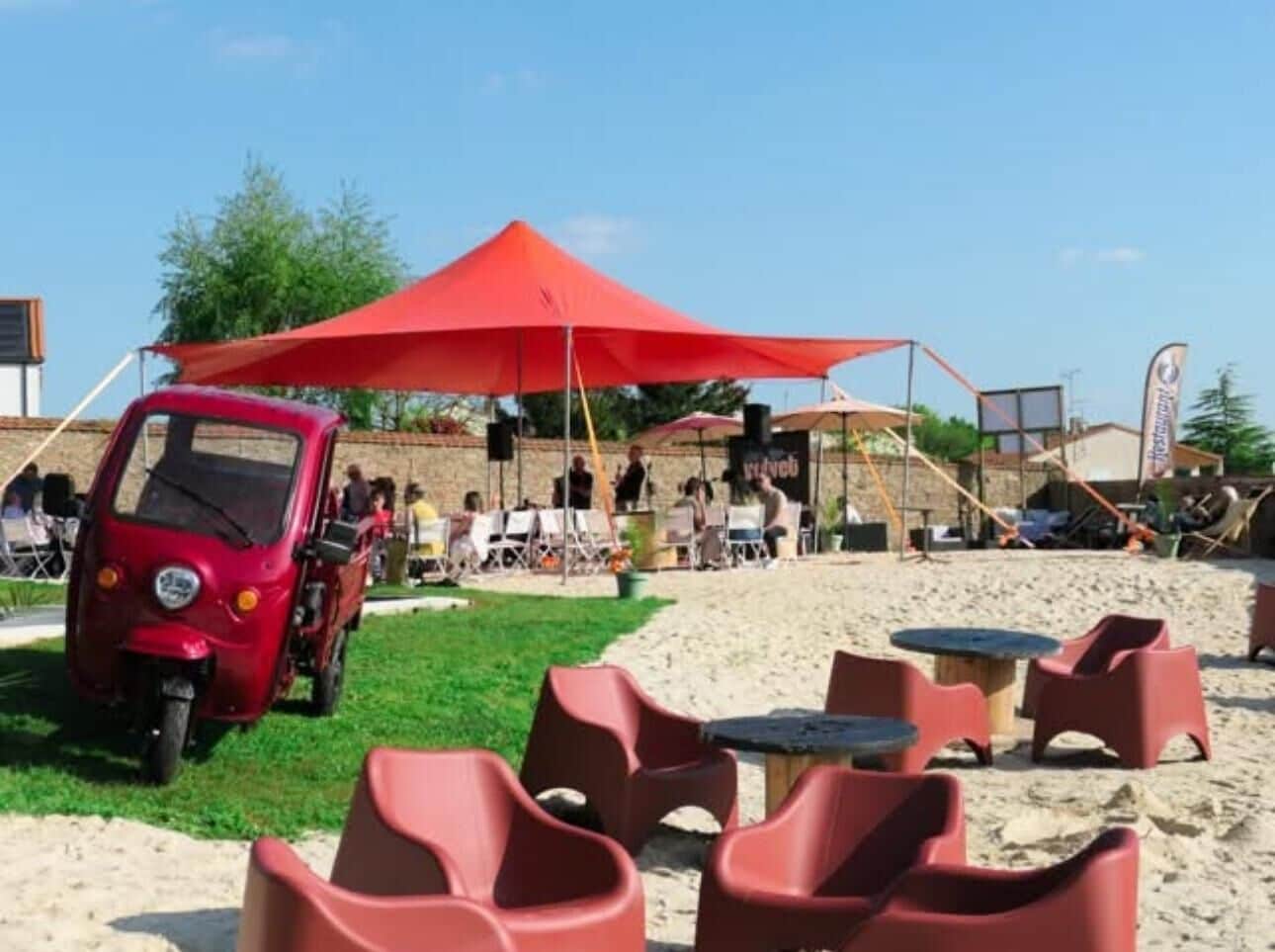 Un décor de plage avec une voiture rouge, des chaises rouges et un grand parasol rouge.