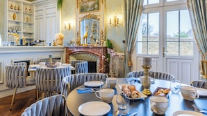 A dining room with a round table set for a meal, a fireplace, and a view through large windows. - Le Château des Tourelles en Vendée (La Gaubretière)