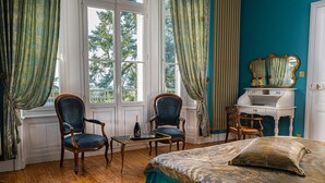 A room with a bed, two armchairs, a small table with a bottle and glasses, a vanity with a mirror, and a window with curtains. - Le Château des Tourelles en Vendée (La Gaubretière)