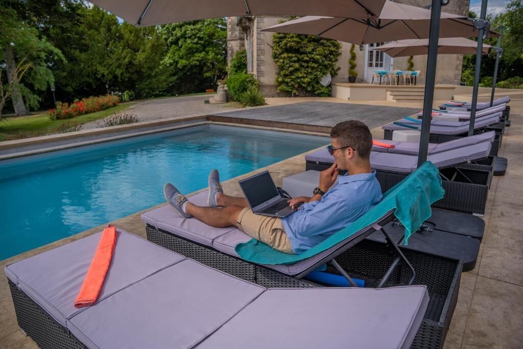Zona junto a la piscina con sillones de descanso y sombrillas, un hombre usando una computadora portátil y una piscina al fondo.