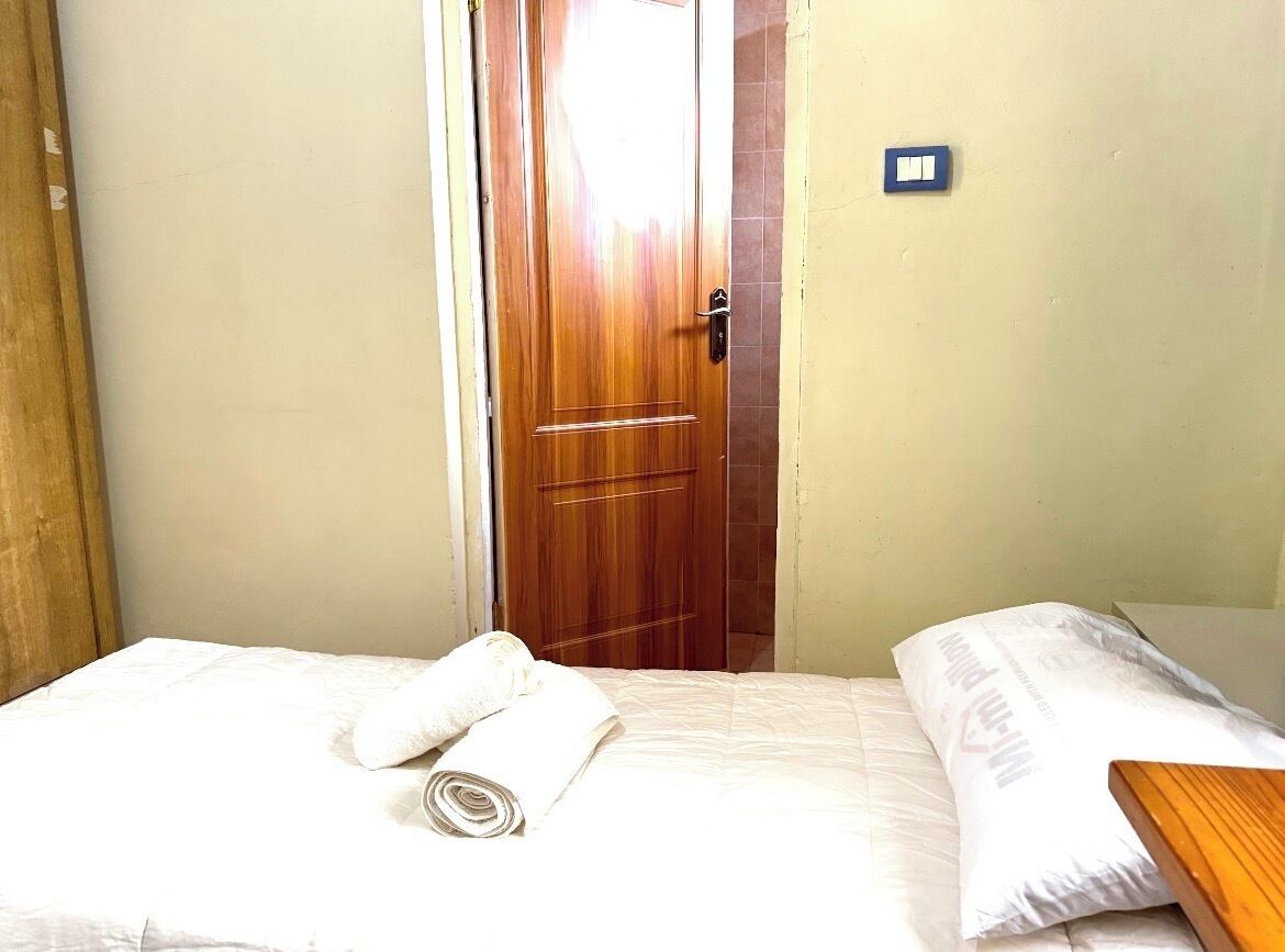 A hotel room with a wooden door, a bed with white bedding, and a small table with a rolled-up towel.