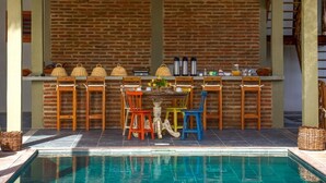 An outdoor bar area with a pool, featuring a brick wall, wooden chairs, and a table with bar stools.