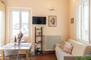 Ein Wohnzimmer mit einer Couch, einem an der Wand befestigten Fernseher, einem Bücherregal und einem Essbereich mit Stühlen und einem Tisch.