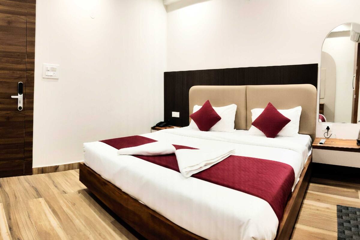 Een hotelkamer met een houten vloer, een bed met wit en rood beddengoed, een zwart hoofdeinde en een nachtkastje met een lamp.