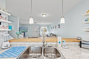 Eine moderne Küche mit einem Spülbecken, einem Mixer und einem Toaster. Es gibt Hängelampen, eine Holz Arbeitsplatte und im Hintergrund ist ein Blick auf das Wohnzimmer.