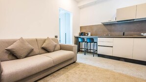 Una sala de estar moderna con un sofá color beige y dos sillones azules, una cocina con armarios blancos y un ventanal grande.