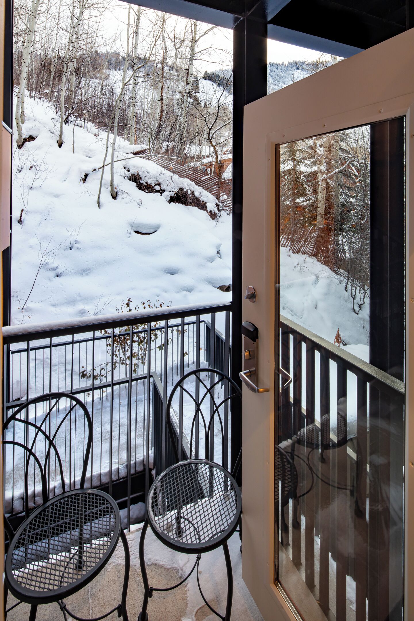 Een balkon met twee metalen stoelen en een glazen deur die uitkomt op een besneeuwde omgeving met bomen.