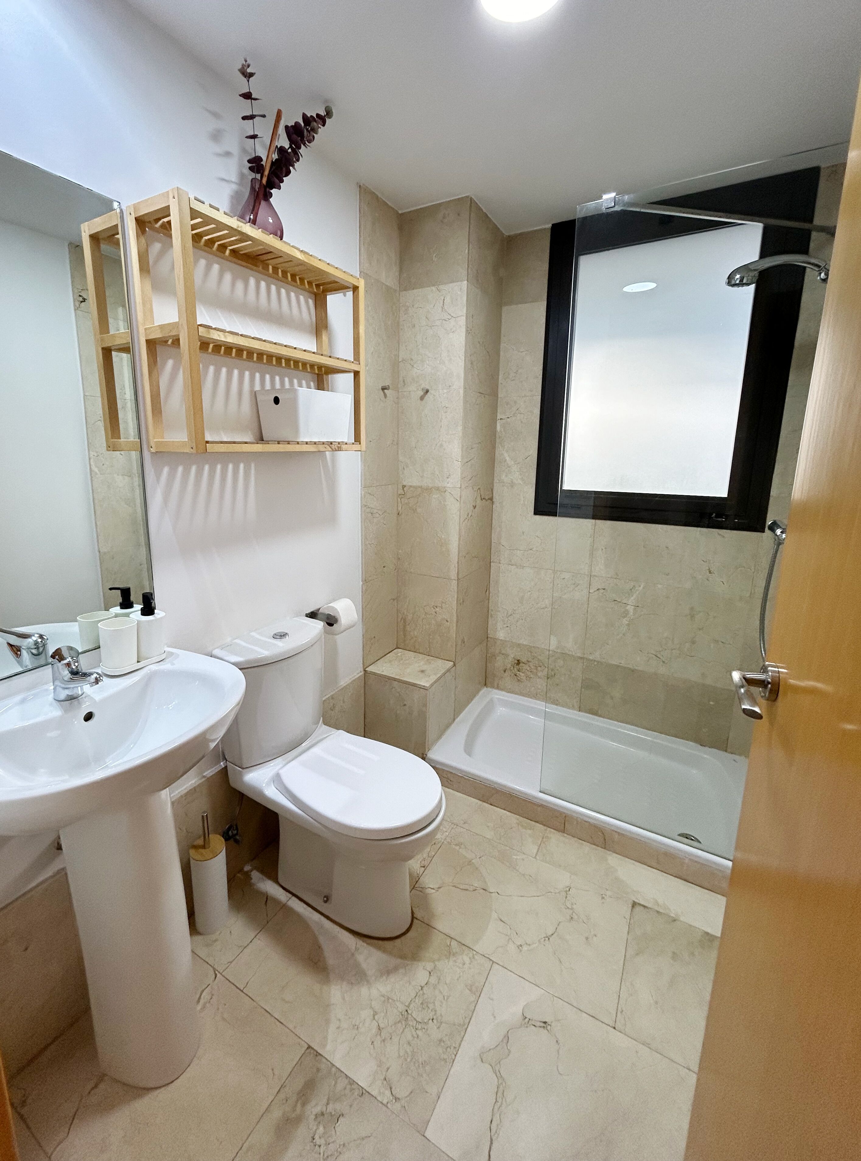 A bathroom with a sink, toilet, and bathtub. There is a wooden shelf above the sink with toiletries.