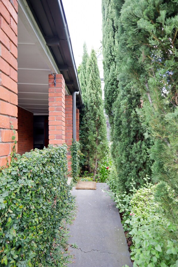 A narrow pathway flanked by dense greenery and a brick building with a visible entrance.