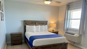 A bedroom with a wooden bed, a nightstand, a lamp, a window with blinds, and a ceiling fan. - Beachgate Condo Suites And Hotel 537a 1 Bedroom Condo (Port Aransas)