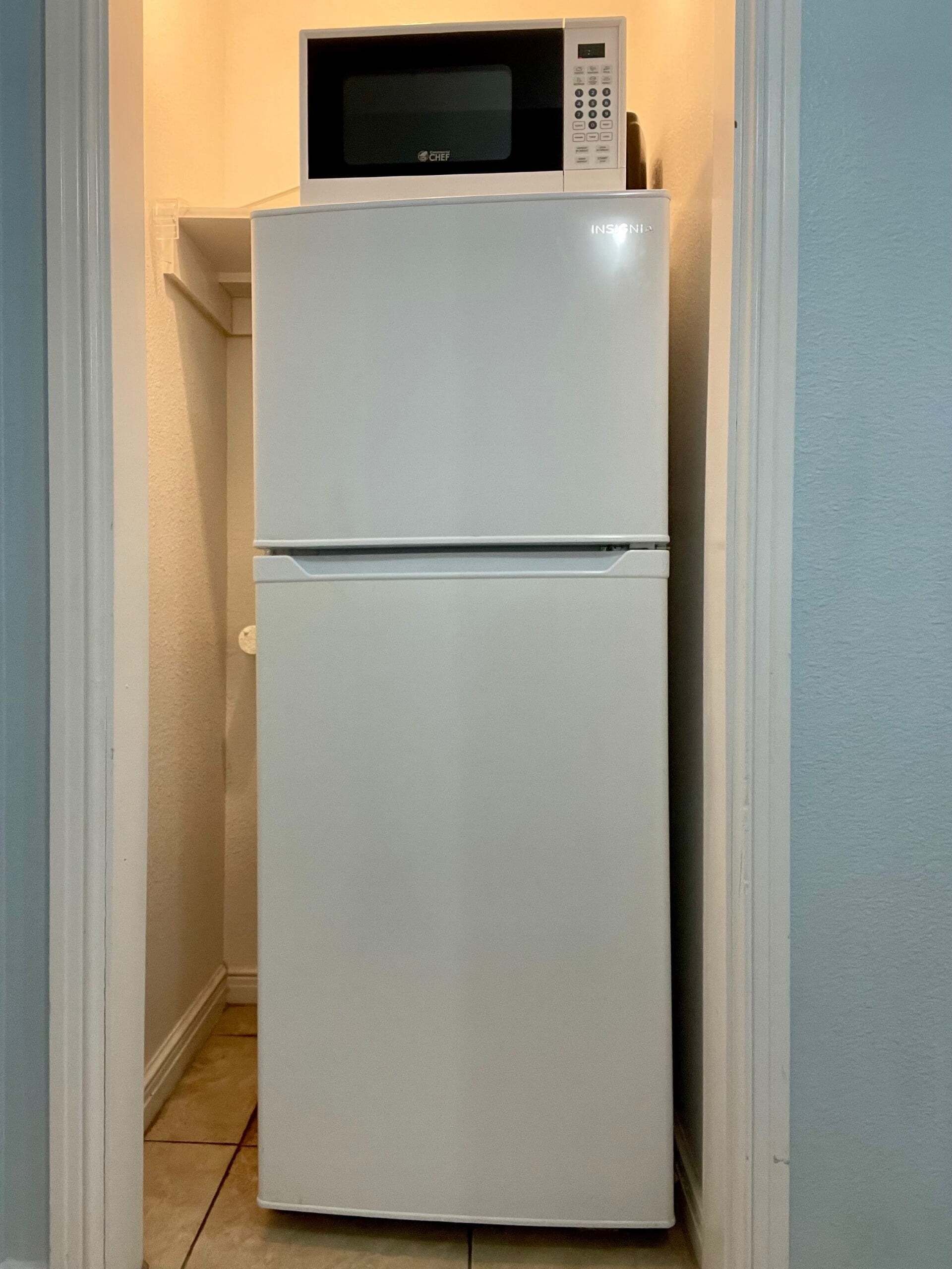 Ein weißer Kühlschrank mit einer Mikrowelle darüber, in einem schmalen Flur mit hellblauen Wänden und Fliesenboden.