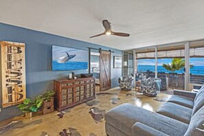 A modern living room with a large TV, wooden cabinet, and a view of the ocean through large windows. - Where Ocean Meets Sky Maalaea Refreshed Oceanfront Condo Panoramic Views (Wailuku)