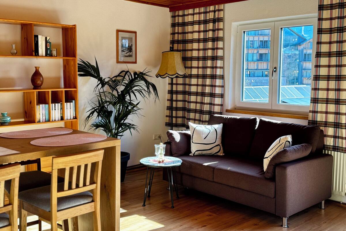 A cozy living room with a wooden ceiling, a sofa, a coffee table, a bookshelf, and a window with plaid curtains.
