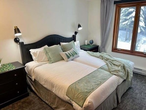 A neatly made bed with a dark headboard, two bedside lamps, and a window with a view of trees.