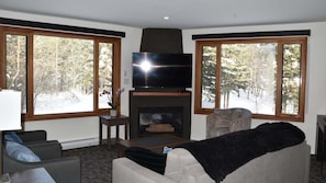 A living room with a fireplace, a television, and a couch. There are two windows with a view of trees and snow.