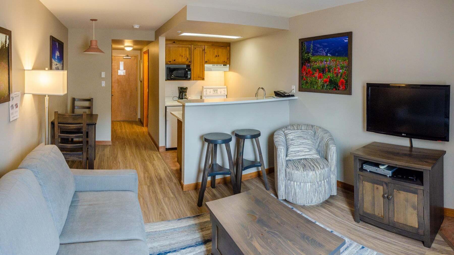 A living room with a sofa, a TV, a wooden cabinet, and a bar stool. There is a kitchen area with wooden cabinets and a microwave.