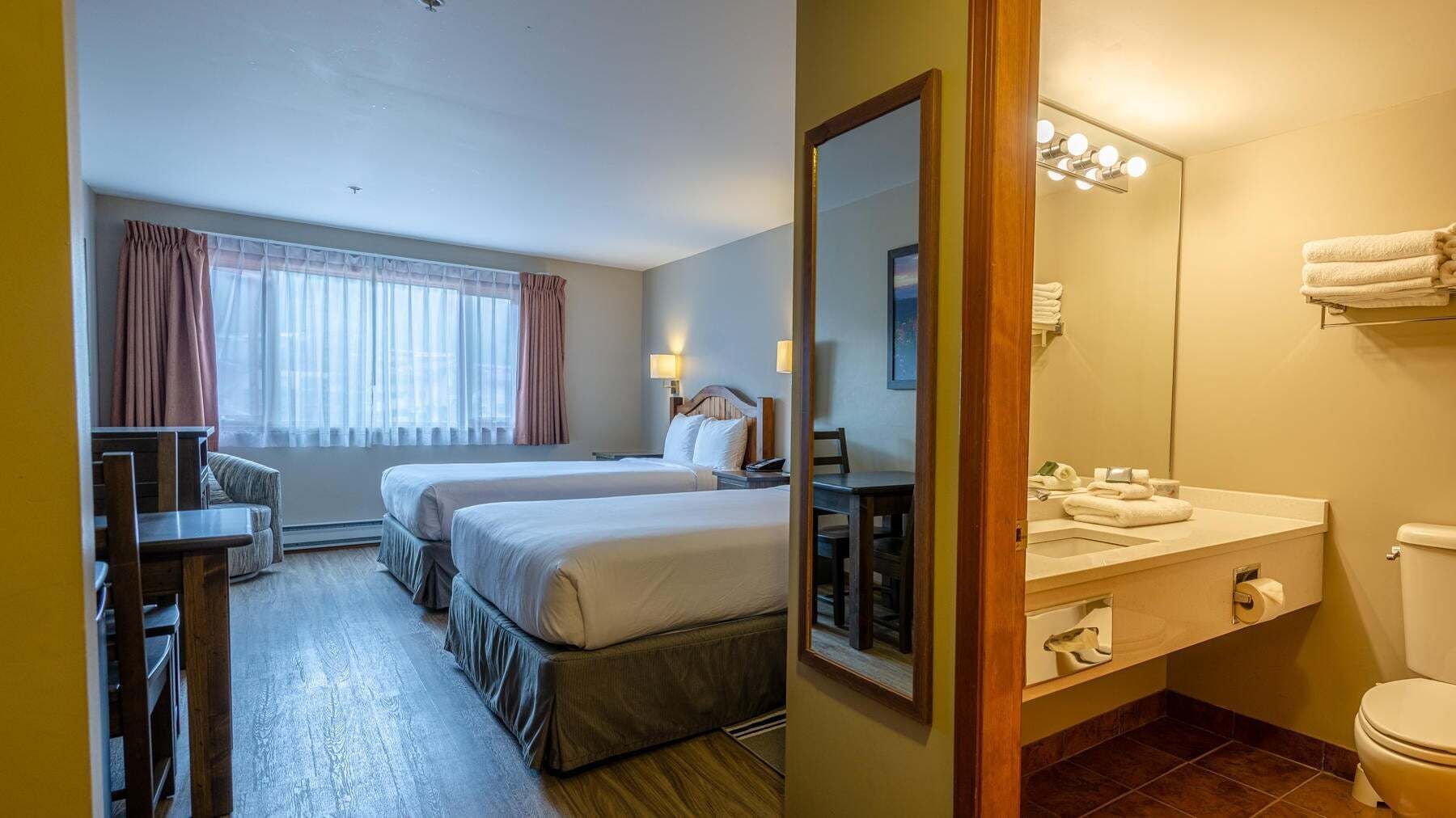 Een hotelkamer met twee bedden, een bureau, een stoel, een badkamer met wastafel en toilet, en een raam met gordijnen.