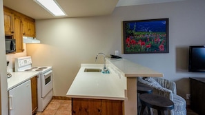 A compact kitchen with white appliances, a wooden cabinet, a sink, and a painting of flowers on the wall. - Cahilty #238/240 1 Bedroom Hotel Room (Sun Peaks)