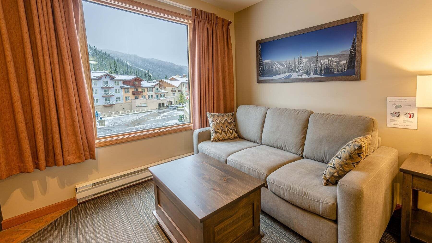 Una sala de estar con un sofá, una mesa de centro de madera y vistas a edificios y colinas nevadas a través de una ventana.