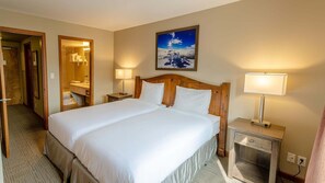 A hotel room with a large bed, two bedside tables with lamps, a bathroom visible through an open door, and a painting of a mountain landscape on the wall.