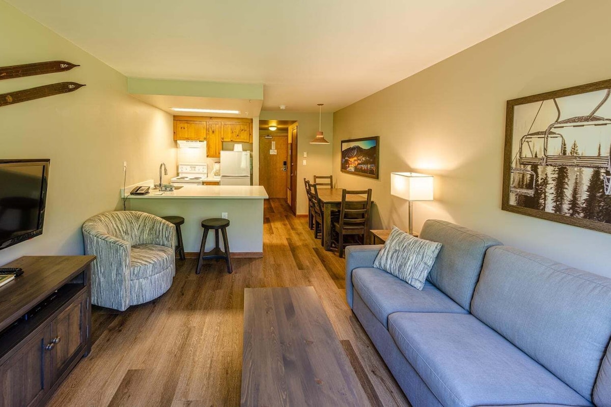 Una sala de estar acogedora con televisor, sofá y sillones. Cuenta con una zona de cocina con armarios de madera y una mesa de comedor.