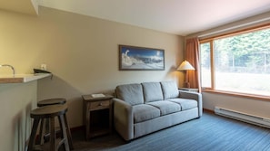 A hotel room with a sofa, a small table, a lamp, a window with a view of trees, and a painting on the wall. - Cahilty #241/243 1 Bedroom Hotel Room (Sun Peaks)
