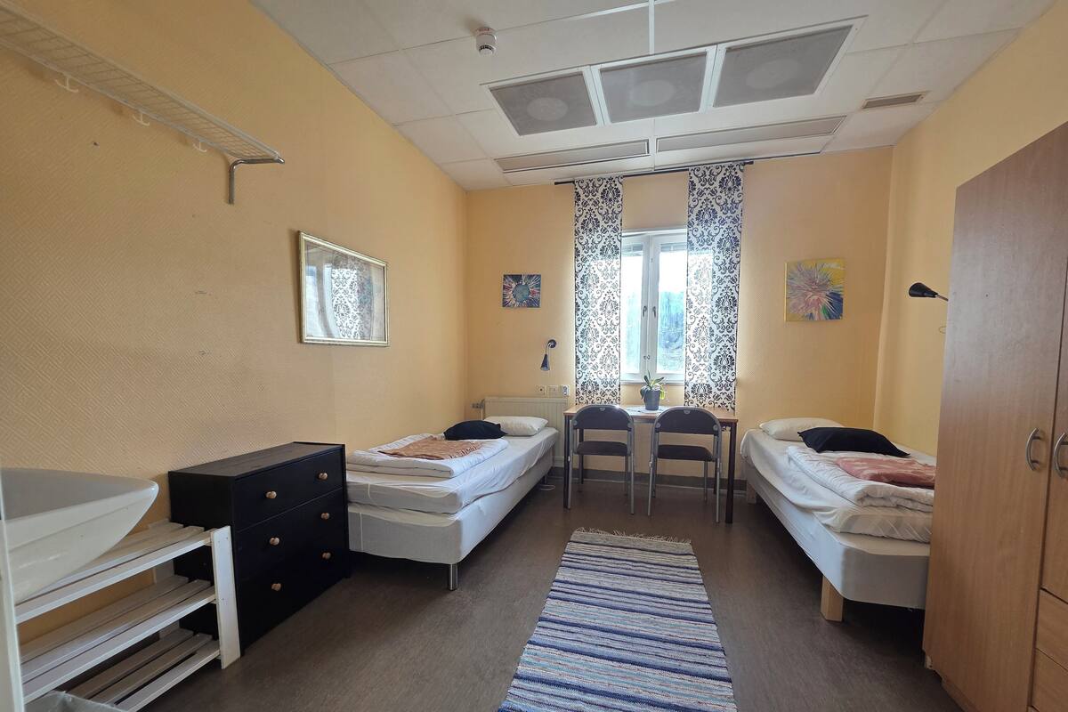 A dormitory room with two single beds, a desk, a chair, a dresser, and a window with curtains.