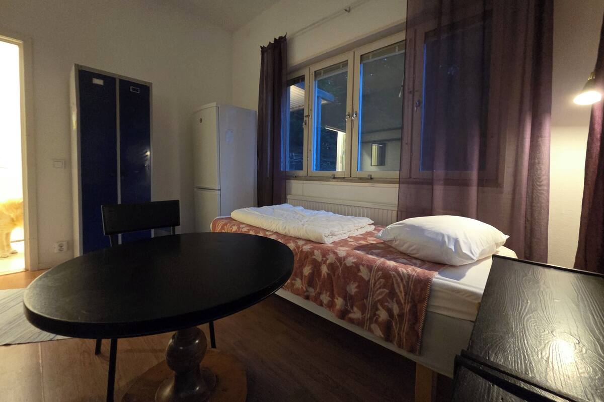 A small, cozy room with a bed, a table, and a chair. There is a window with curtains, a door, and a radiator under the window.